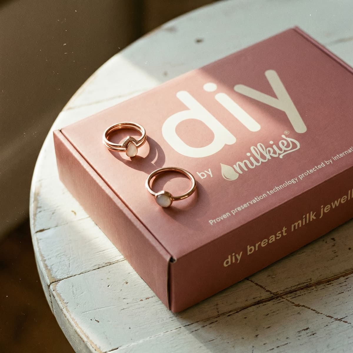 Two rose-gold DIY by MILKIES at-home kit pieces featuring diy breastmilk rings with milky white stones resting on a pink breast milk jewelry kit box.