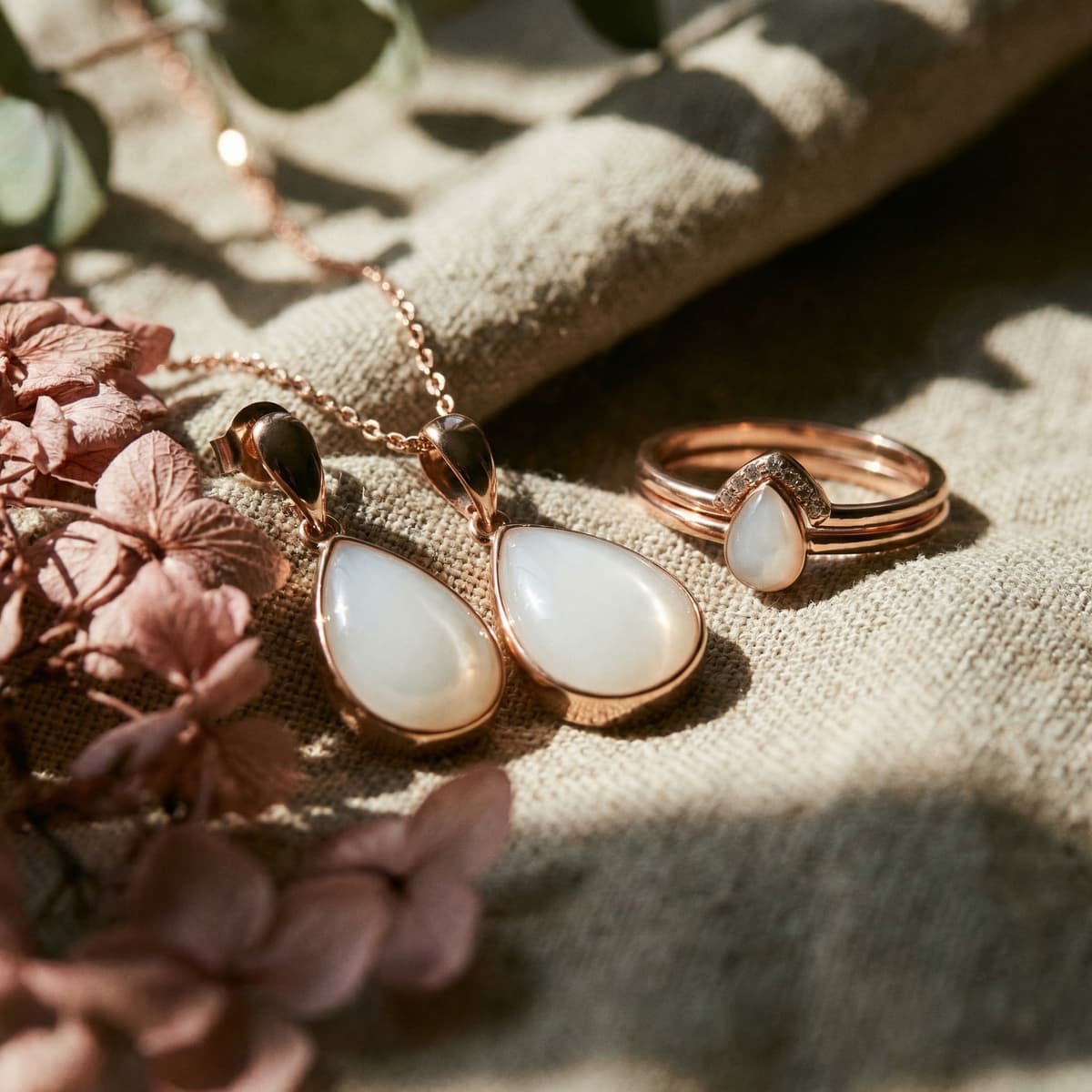 Rose gold breastmilk jewelry set for moms drying up milk, featuring a teardrop pendant necklace, matching earrings, and a stacking ring with a milky white resin stone on soft linen with blush flowers.