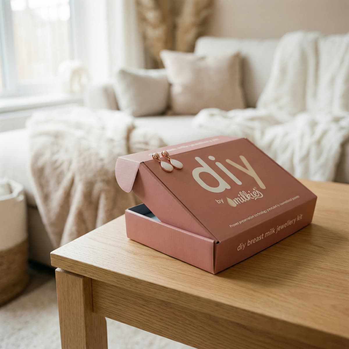 DIY by MILKIES breast milk jewellery kit in a pink box on a wooden table in a cozy living room, an end of breastfeeding gift for making a keepsake necklace, ring, or earrings at home.