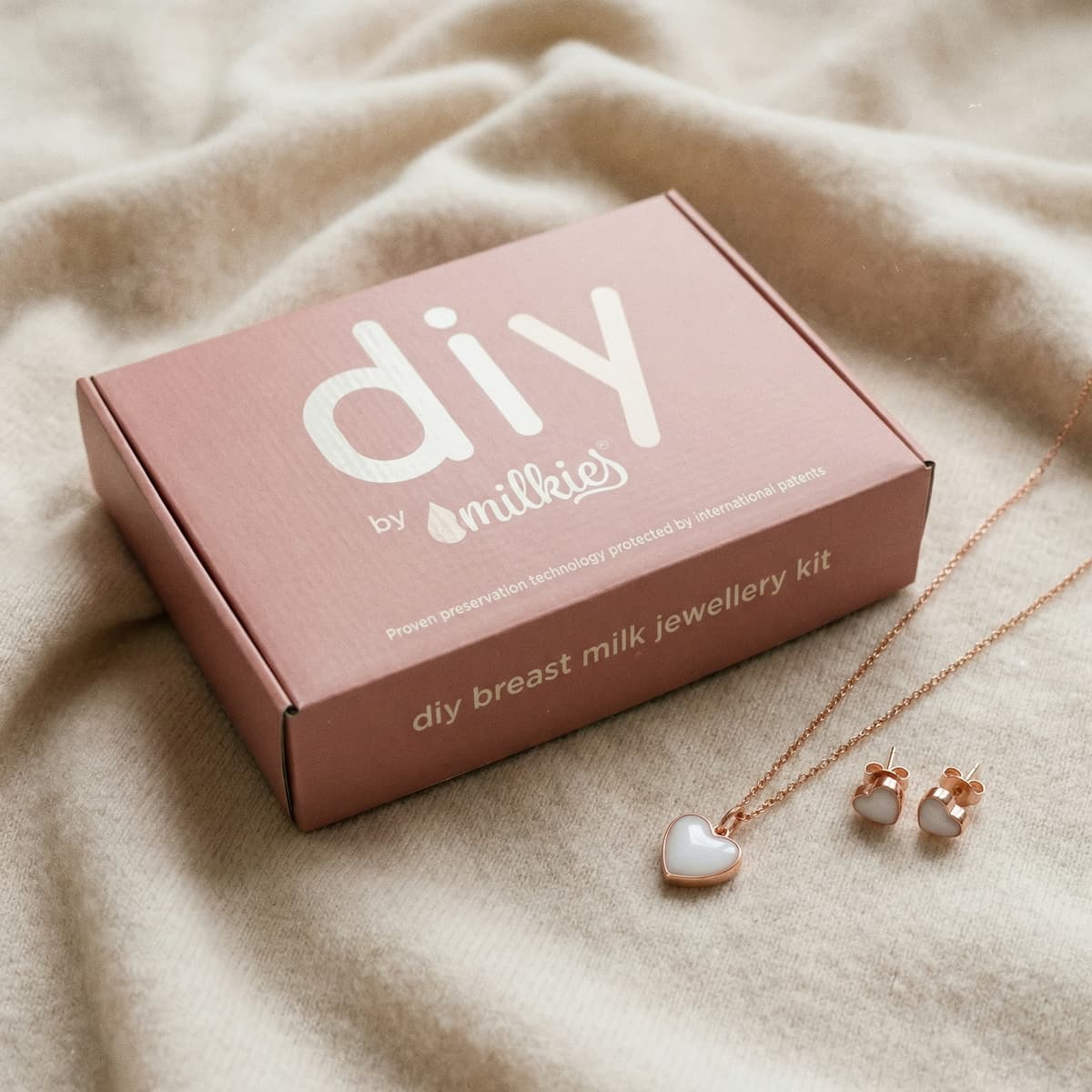 Pink DIY by MILKIES breast milk jewelry kit box on soft fabric with rose-gold heart necklace and matching heart earrings, a meaningful gift for new mother keepsake