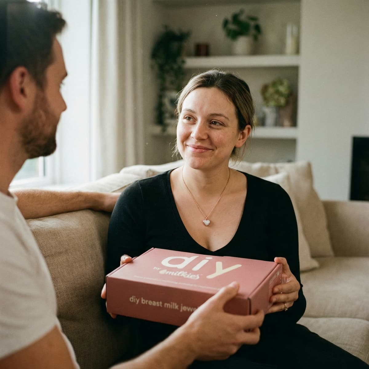 Partner hands a new mom a DIY by MILKIES at-home breastmilk jewelry kit box on a cozy sofa, a meaningful gift for new mother and an easy way to create a personal keepsake at home.