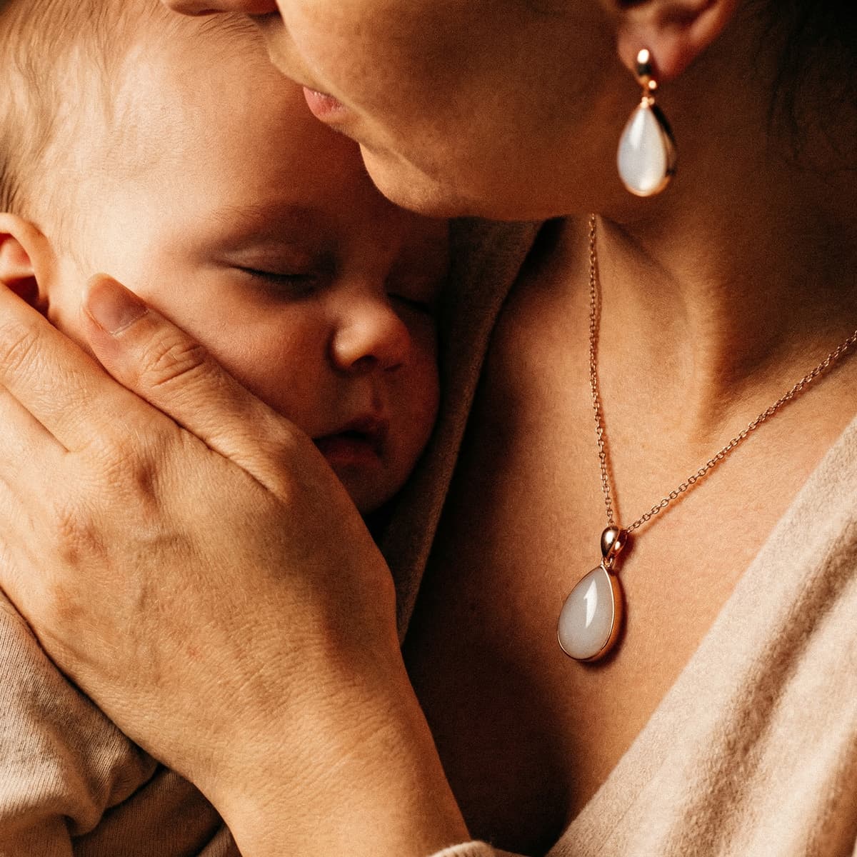 Mother cuddling a sleeping newborn while wearing golden boobs breastmilk keepsake jewelry, featuring a gold-plated teardrop pendant necklace and matching earrings.