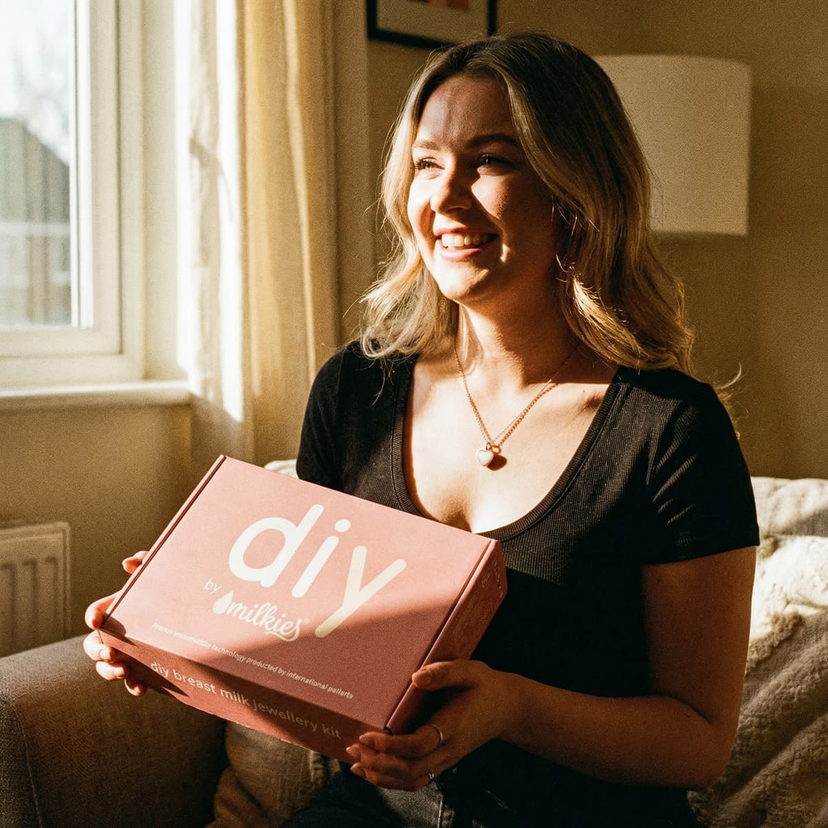 Smiling woman holding a DIY by MILKIES breast milk jewellery kit box in warm window light, explaining why making golden boobs keepsakes at home is a meaningful, private, and convenient choice.