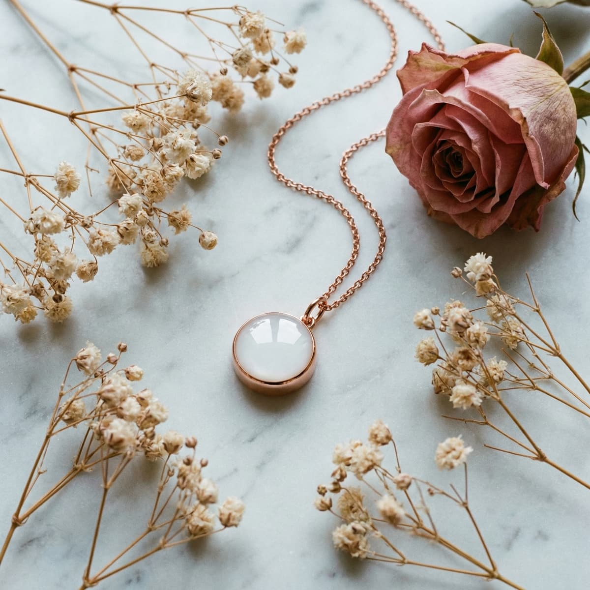 Rose gold necklace featuring a round white resin pendant milk keepsake, photographed on marble with dried baby’s breath and a blush rose.