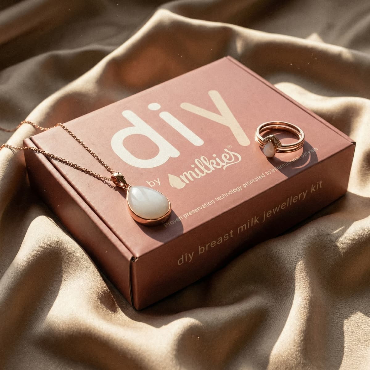 Rose-gold DIY by MILKIES breast milk jewelry kit box featuring a necklace pendant and matching ring on soft fabric, a keepsake idea for new mom gifts