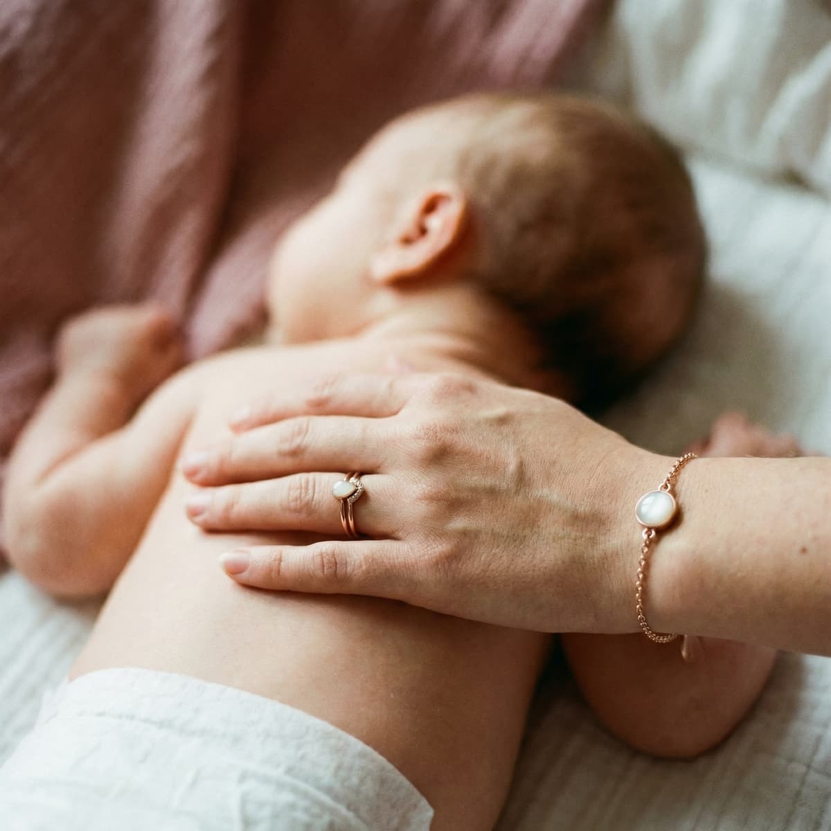 Mother gently holds a sleeping newborn while wearing a rose-gold ring and bracelet featuring milky white stones, a nursing keepsake jewelry set.