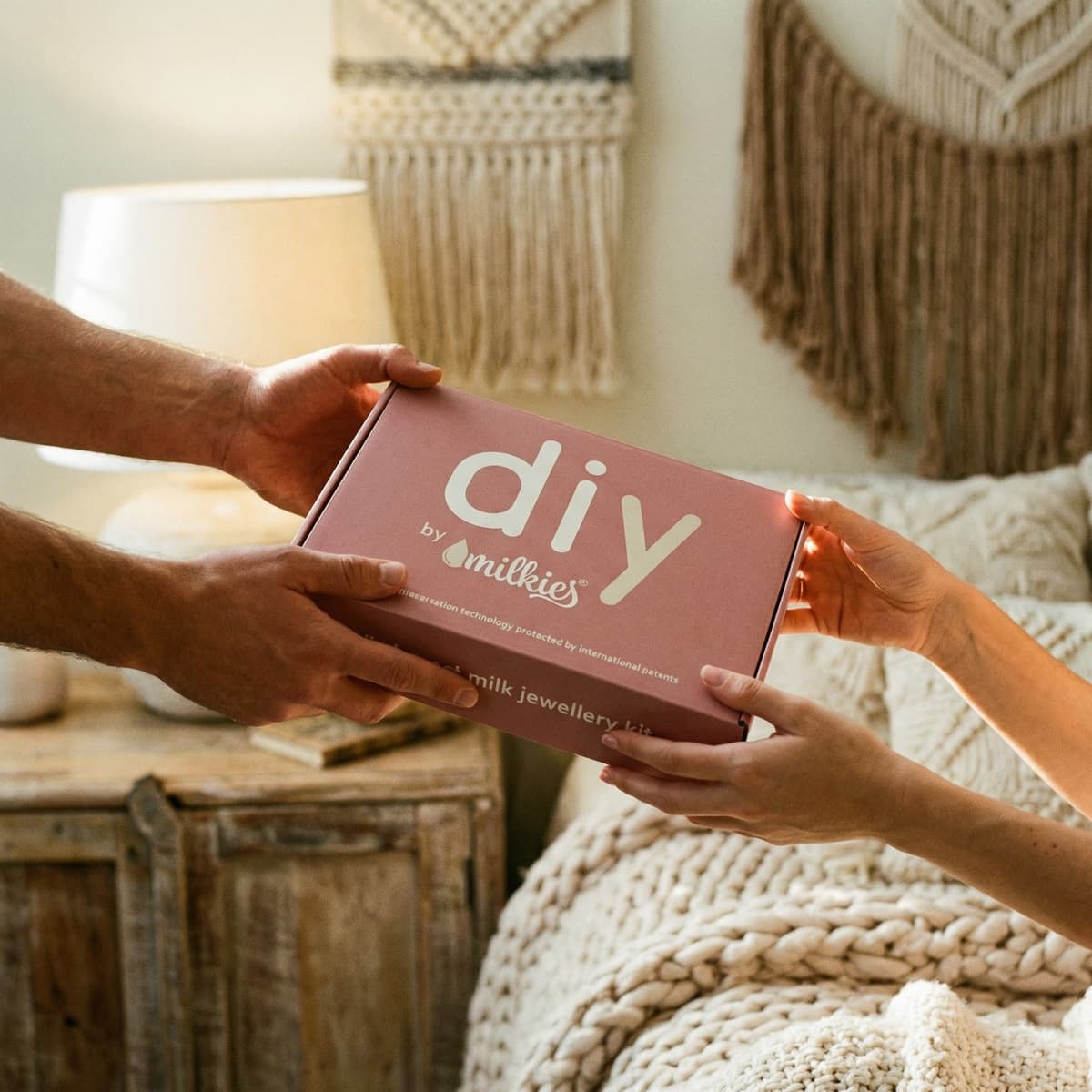 Hands exchange a pink DIY by MILKIES breastmilk jewellery kit box as a thoughtful push present in a cozy bedroom setting