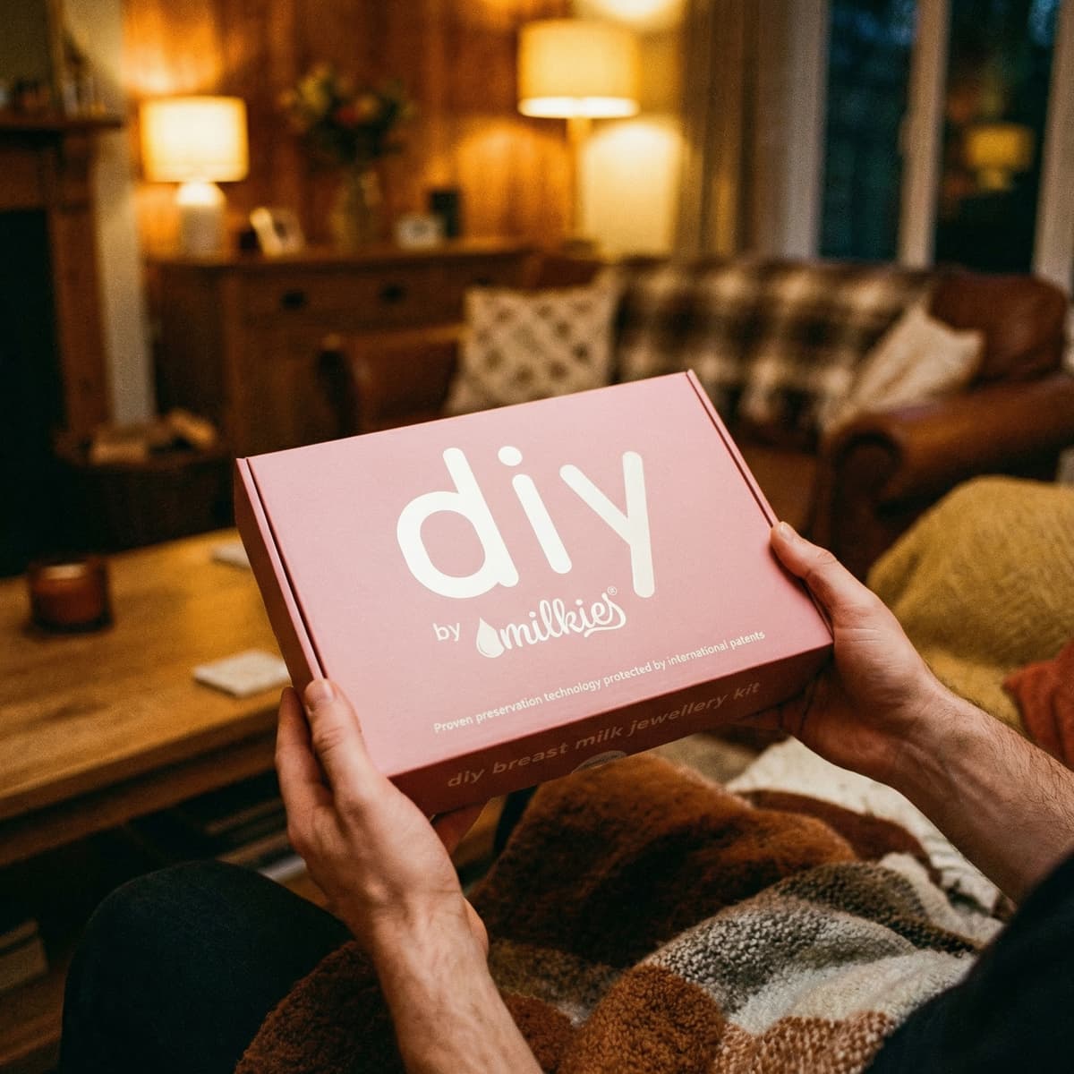 Hands holding a DIY by MILKIES breast milk jewellery kit box in a cozy living room, an at-home keepsake-making push present for wife with patented preservation technology.