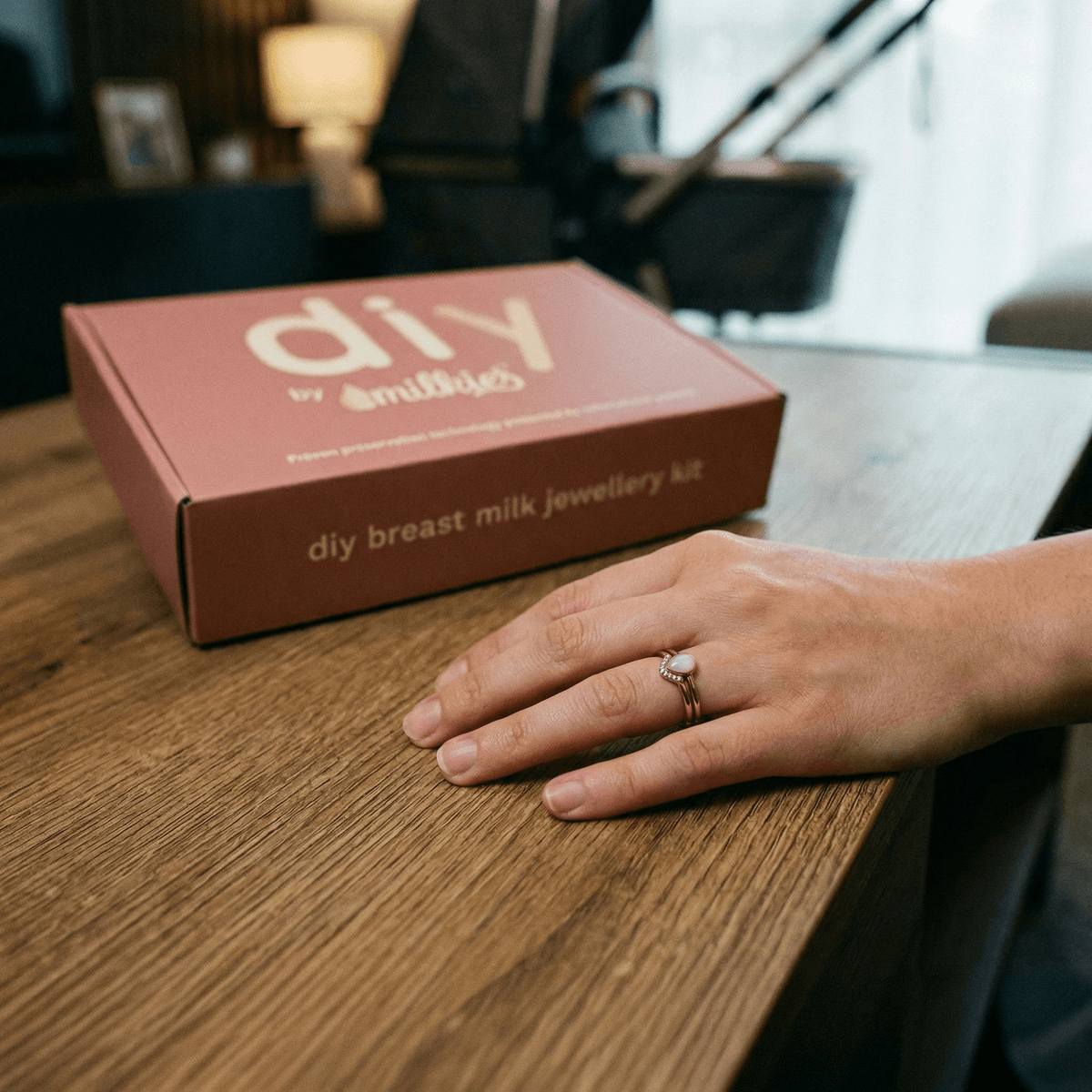 Hand resting on a wooden table wearing a breastmilk keepsake ring next to a DIY by MILKIES breast milk jewelry kit box, highlighting unique push presents for new moms.