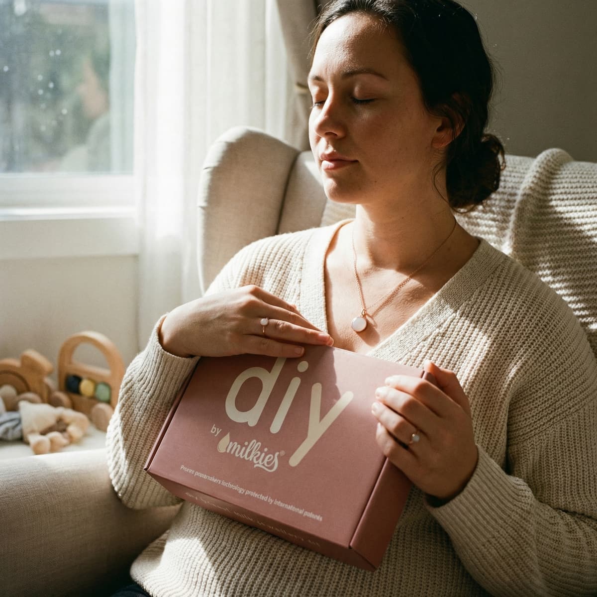 A relaxed mom in a sunlit chair holding a pink DIY by MILKIES keepsake kit box, highlighting an at-home option for creating unique push presents with a meaningful breastmilk jewelry kit.