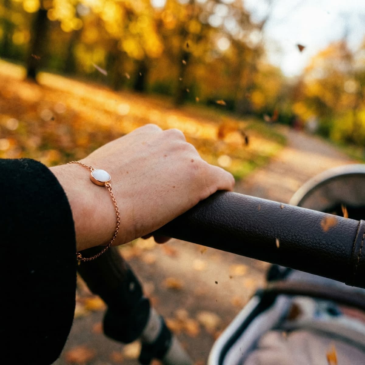 Hand pushing a stroller along an autumn path, wearing a rose-gold bracelet with a white oval stone as a 1 year breastfeeding keepsake jewelry piece.