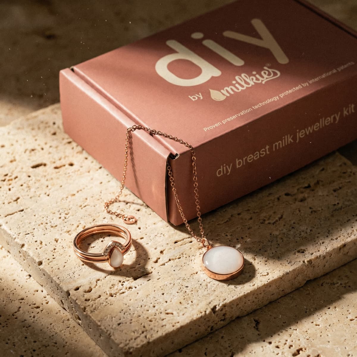 Rose-gold DIY by MILKIES box displaying a breast milk jewellery making kit with ring and necklace featuring white keepsake stones on a textured stone surface.