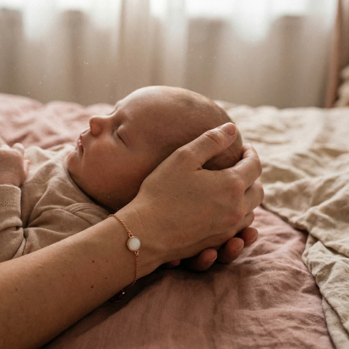 Mother cradling a sleeping newborn on soft bedding while wearing a delicate keepsake bracelet, showing why a breastmilk bracelet diy making kit is a meaningful at-home way to preserve the bonding moment.