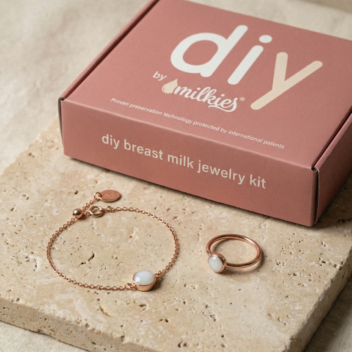 DIY by MILKIES breastmilk bracelet making kit displayed with a pink “DIY breast milk jewelry kit” box next to a rose-gold bracelet and ring featuring milky white resin stones on a neutral stone surface.