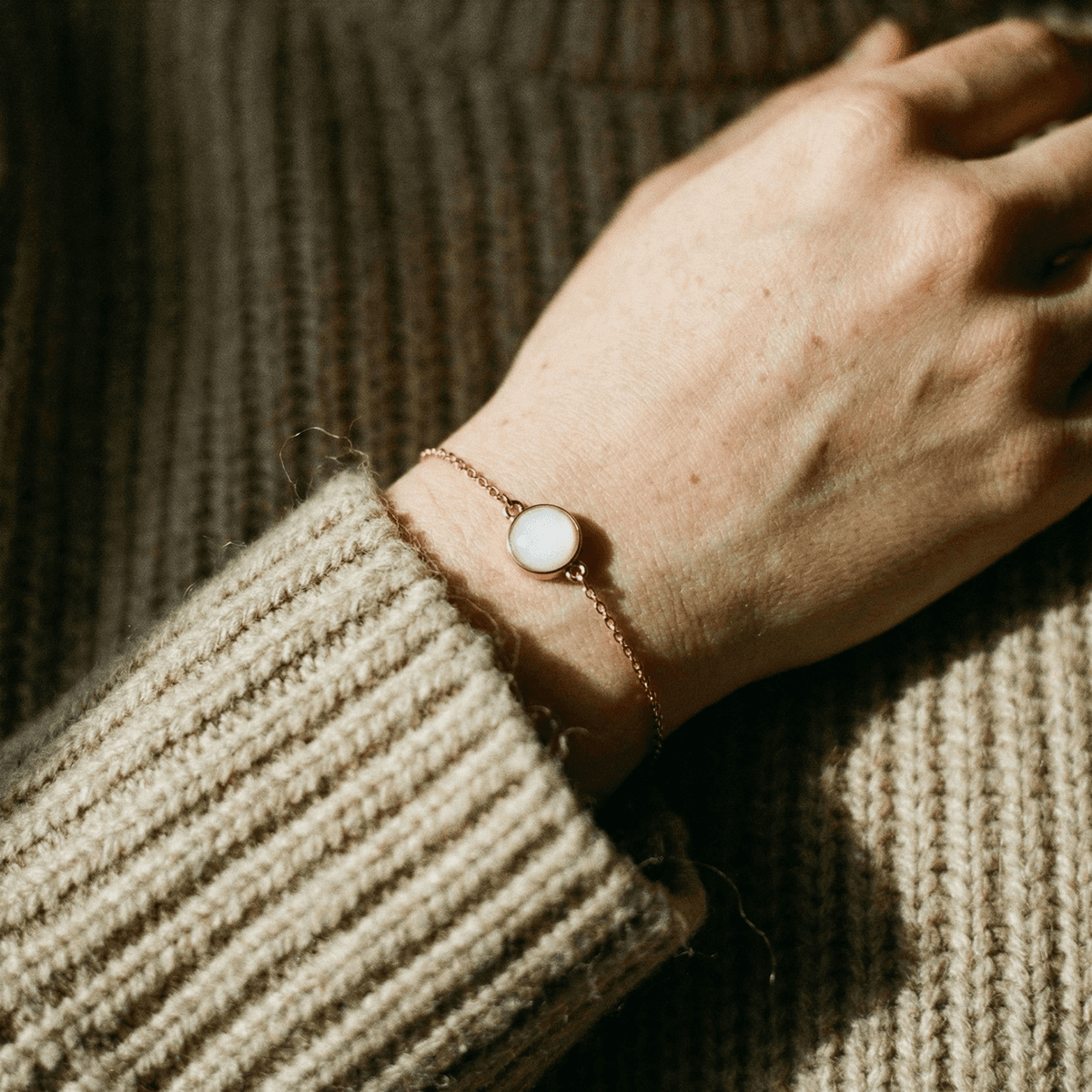 Close-up of a hand wearing a rose-gold bracelet with a milky white resin charm, showing finished results made with a breastmilk bracelet making kit against a cozy knit sweater background.