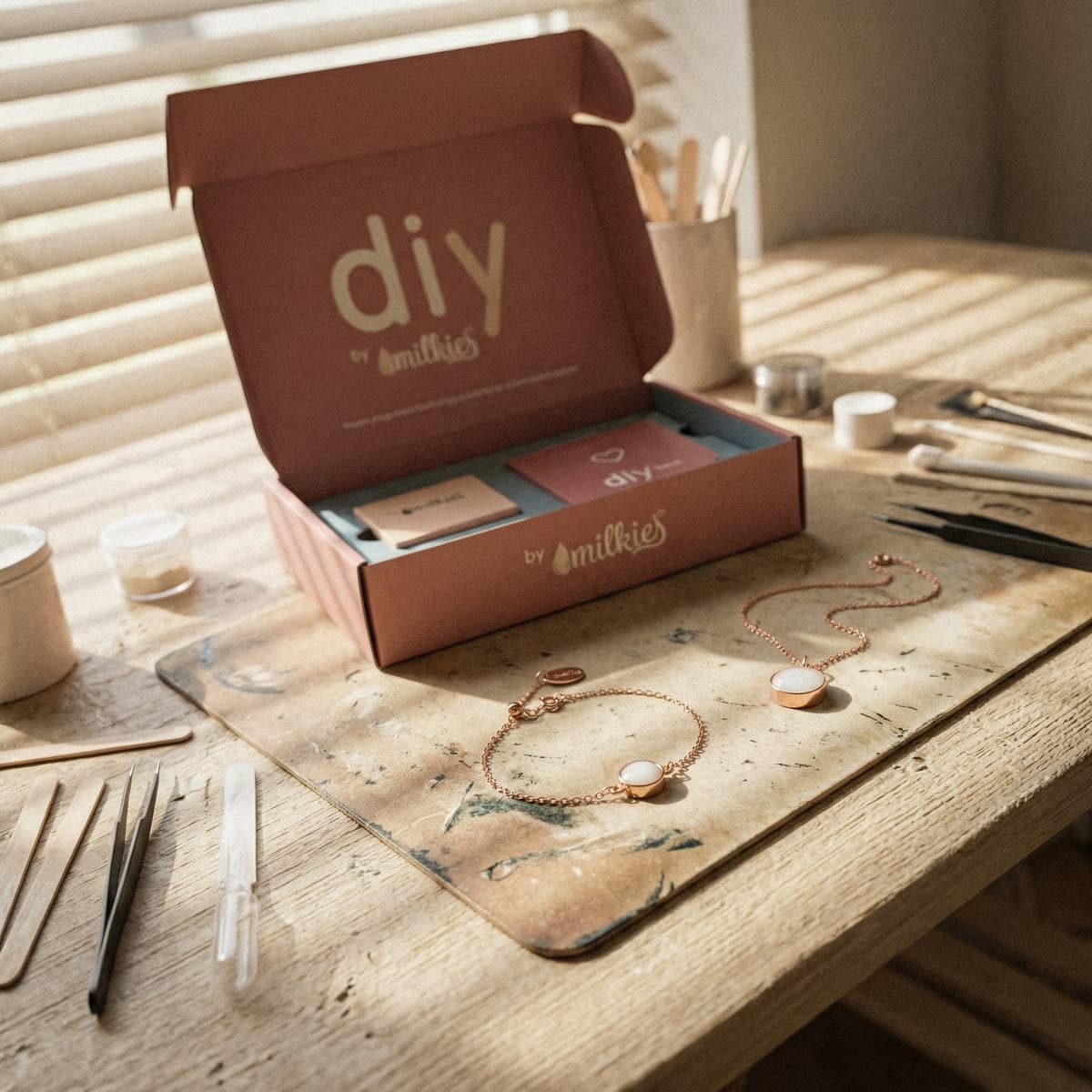 Open DIY by MILKIES at-home jewelry workshop box on a wooden table in warm sunlight, showing tools, resin supplies, and rose-gold bracelet and necklace settings for a breastmilk bracelets diy making kit.