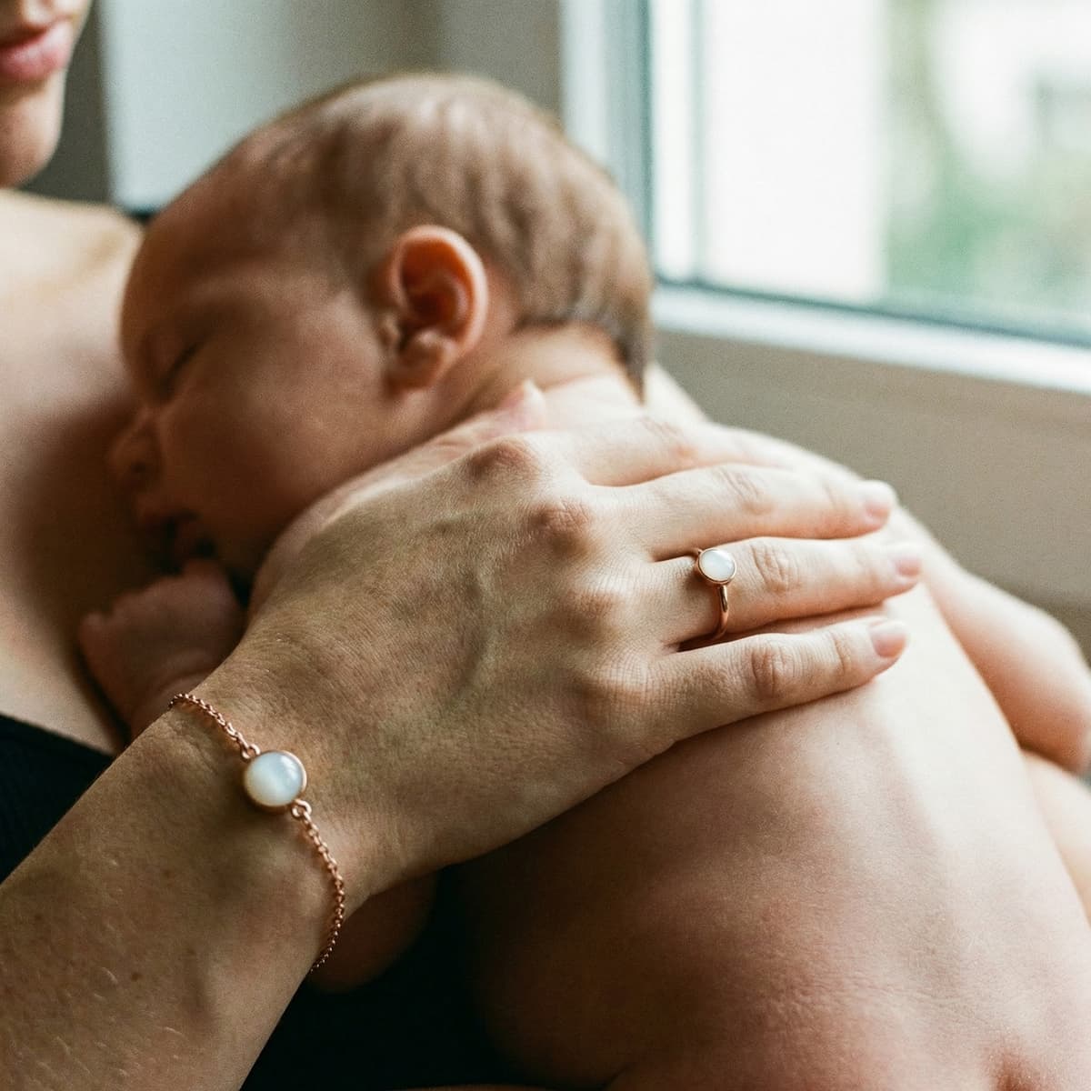 Mother holding a sleeping newborn by a window, wearing a breastmilk bracelet and ring, showing why a breastmilk bracelets diy making kit is a meaningful at-home way to create a personal keepsake.