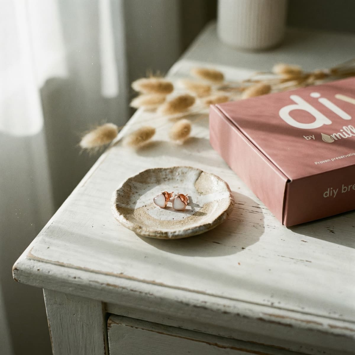 Rose-gold heart stud breastmilk earrings displayed on a ceramic dish beside DIY by MILKIES packaging from a breastmilk earring making kit on a white bedside table in sunlight.
