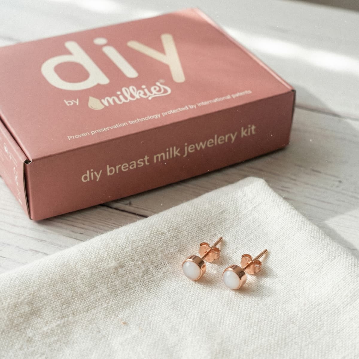 Rose-gold stud earrings with milky-white resin stones on a linen cloth beside a DIY by MILKIES box, showing an at-home breastmilk earrings making kit for creating breast milk jewelry.