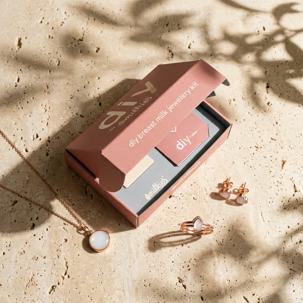 Blush pink DIY by MILKIES box with a breastmilk jewellery making kit displayed on a textured stone surface, alongside matching keepsake necklace, ring, and earrings in soft shadowed light.
