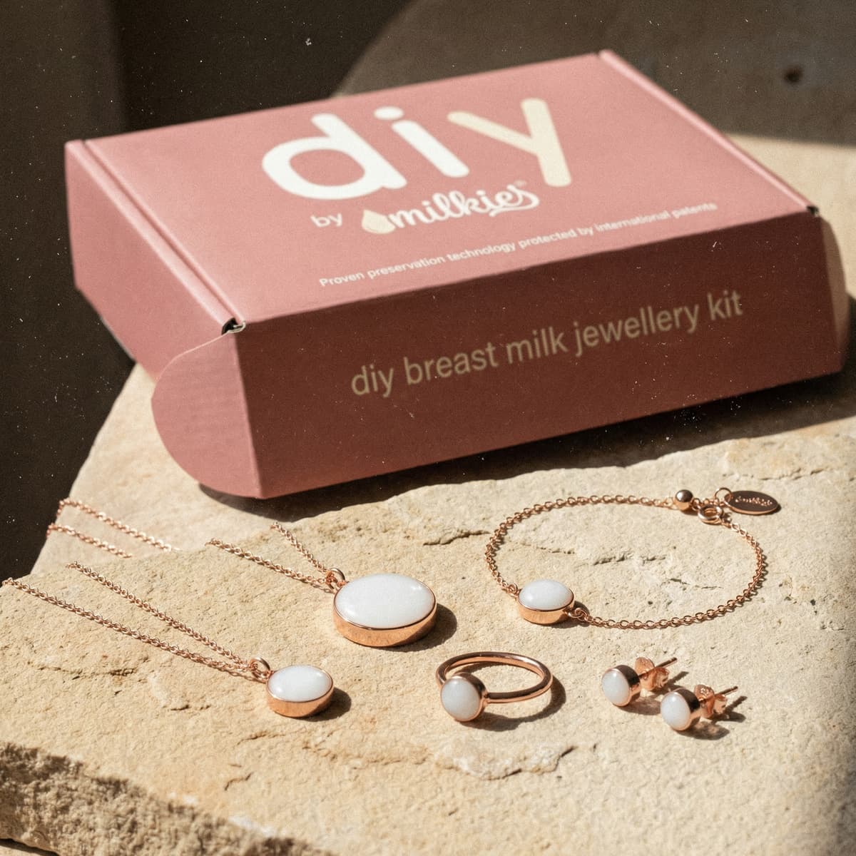 Rose-pink DIY by MILKIES box and finished keepsakes displayed as a breastmilk jewelry diy making kit, featuring rose-gold necklace pendants, bracelet, ring, and stud earrings with milky-white resin stones on a stone surface.