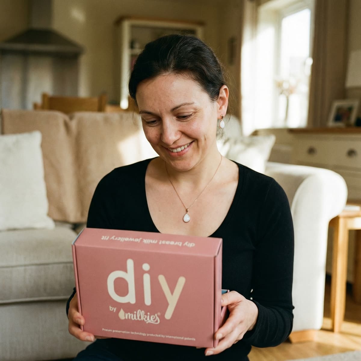 Smiling mom at home holding a pink DIY by MILKIES box, highlighting why this breastmilk necklaces diy making kit is a convenient at-home option for creating a personal keepsake with patented preservation technology.