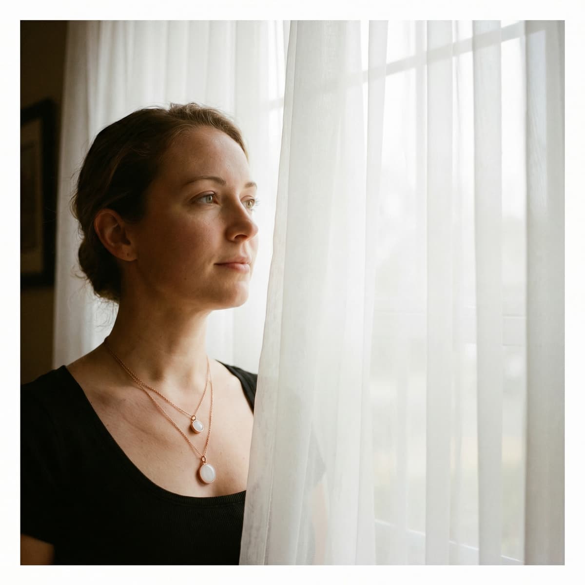 Woman wearing two delicate breastmilk necklaces made with an at-home breastmilk necklaces making kit, standing by a bright window with sheer curtains.