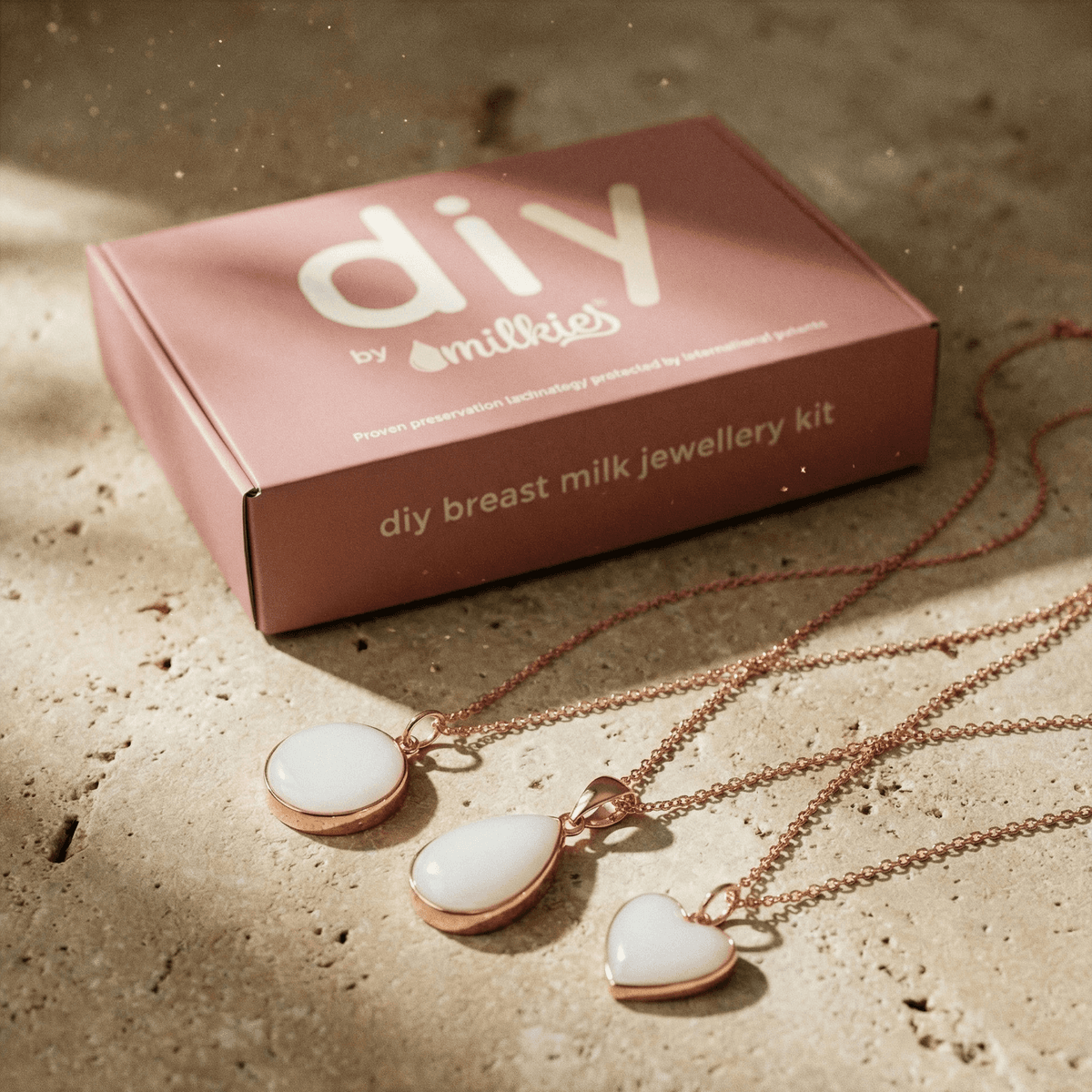 Pink “DIY breast milk jewellery kit” box from DIY by MILKIES next to three rose-gold breastmilk necklaces on a stone surface, showcasing a breastmilk necklaces making kit with white resin pendants