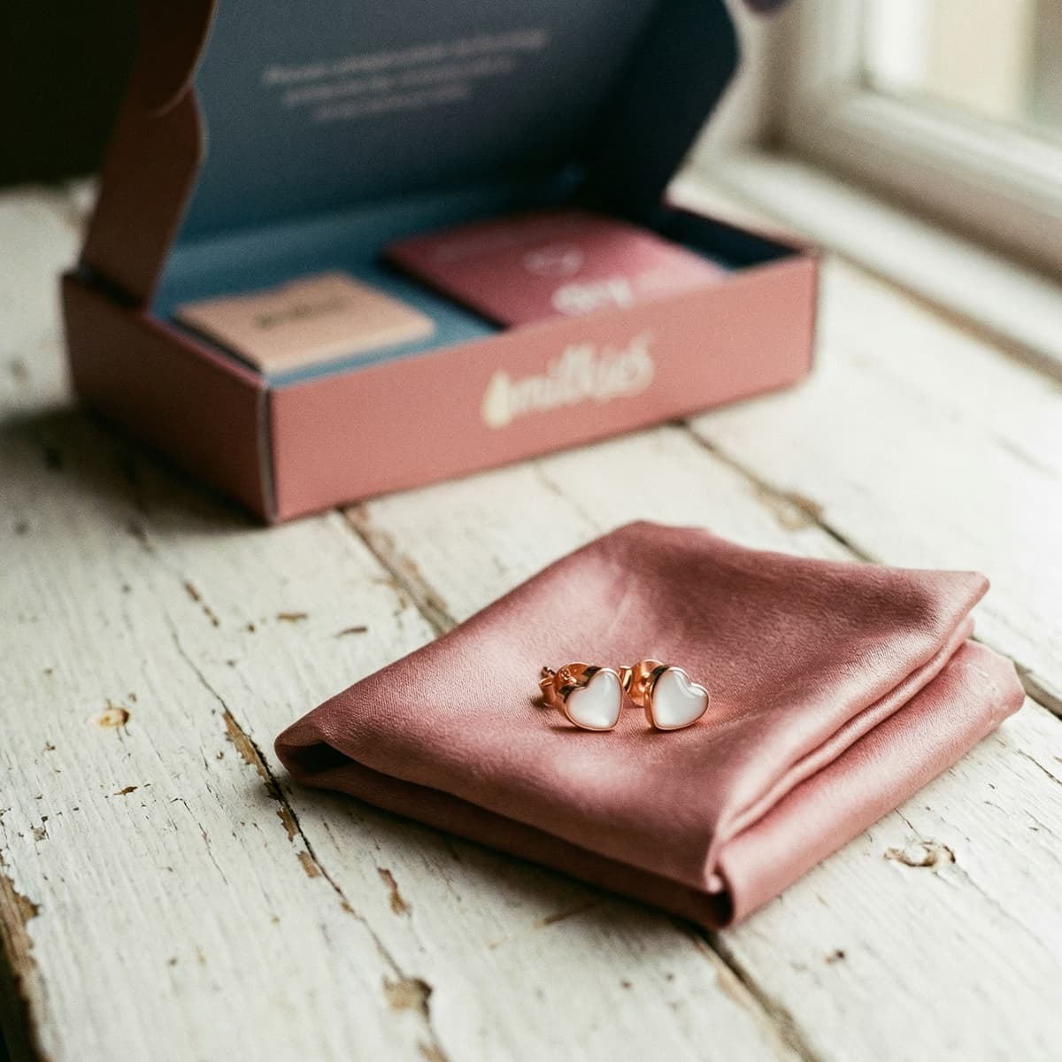 Rose-gold heart stud earrings on a blush cloth next to a branded box, showcasing a diy breastmilk earring making kit for creating keepsake jewelry at home.
