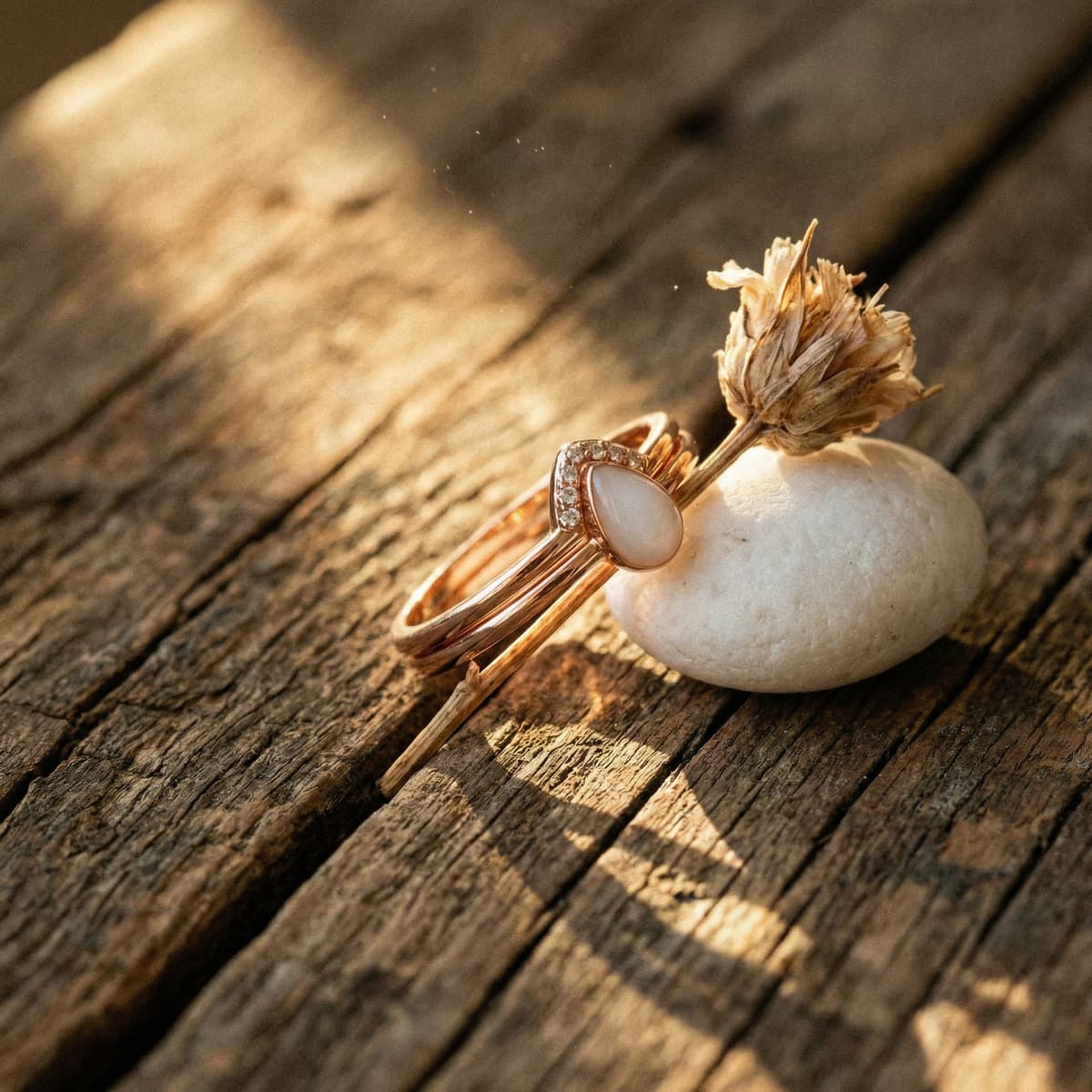 Rose gold teardrop breastmilk stone ring with pavé accents, finished using a diy breastmilk ring making kit, displayed on rustic wooden boards beside a white pebble and dried flower.