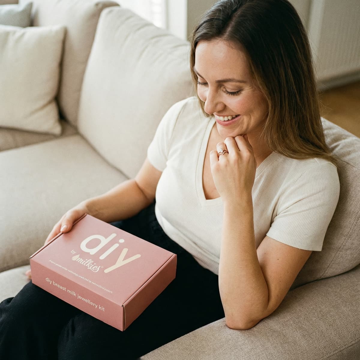 Smiling woman on a sofa holding a DIY by MILKIES box, highlighting why an at-home diy breastmilk ring making kit is a convenient way to create a meaningful keepsake.