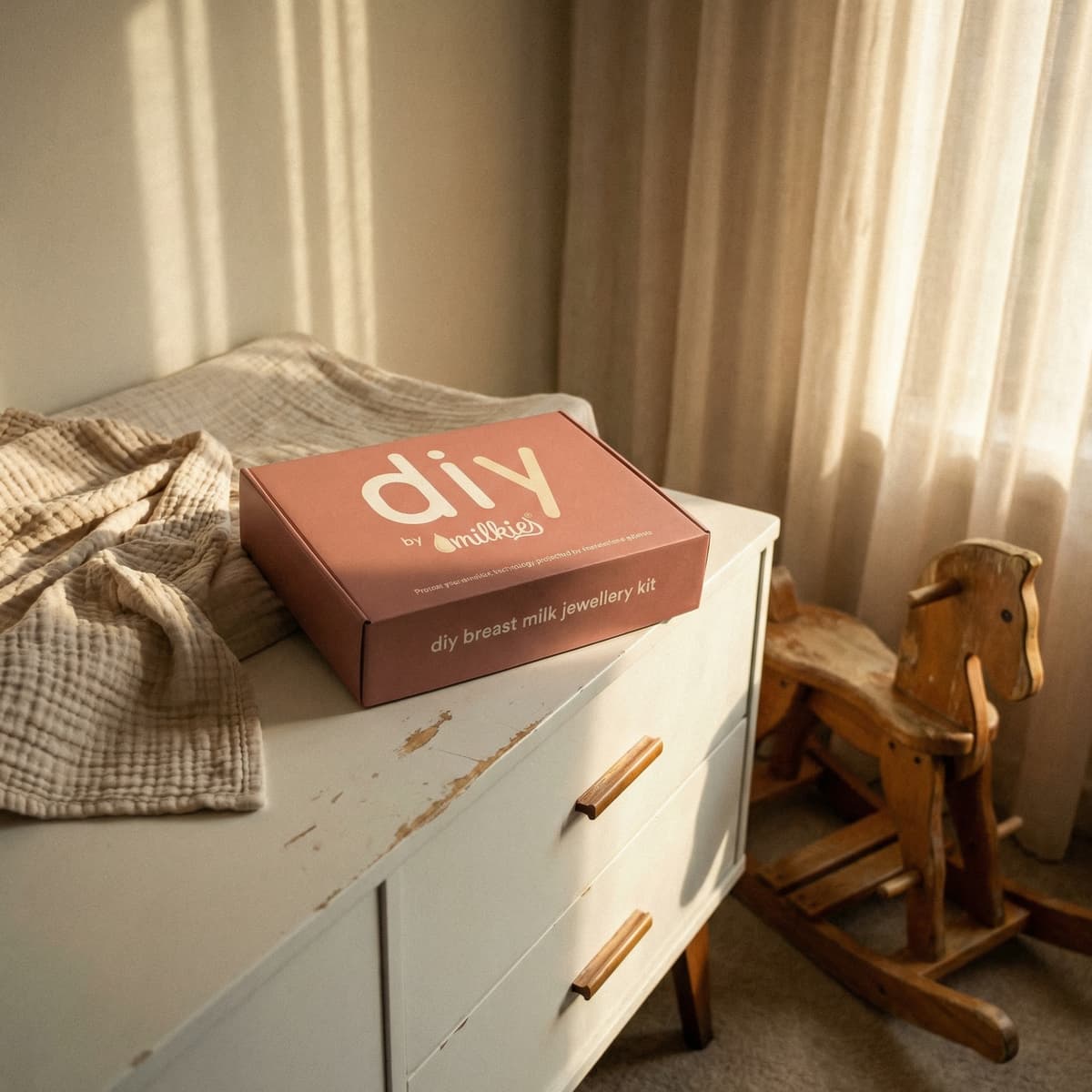 Sunlit nursery dresser scene with DIY by MILKIES breast milk jewelry kit box, baby blanket, and wooden rocking horse, perfect for new mom gifts for pregnant women.