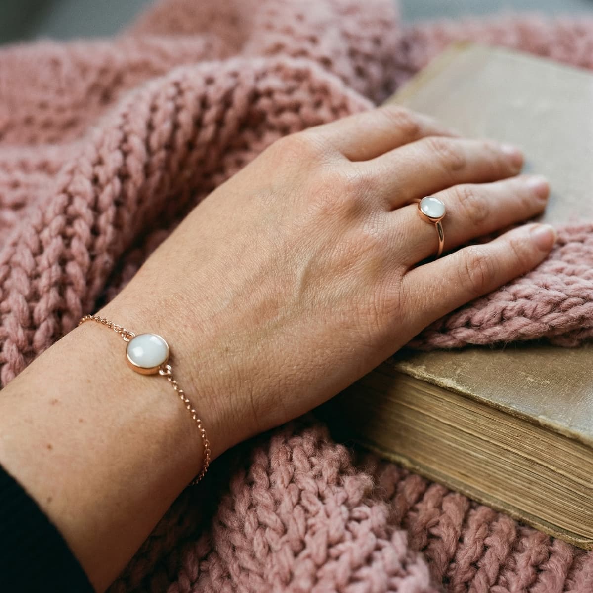 Hand wearing a delicate gold ring and matching bracelet with milky white stones, a stopping breastfeeding keepsake jewelry set resting on a pink knitted blanket beside a vintage book.